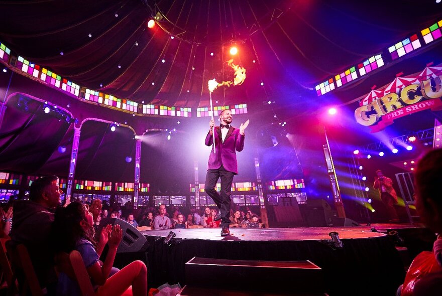 The ringmaster of the circus on a big top stage, holding a firestick, surrounded by a seated audience.