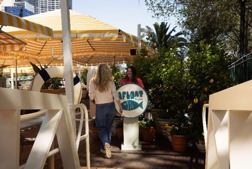 Person walking past a receptionist standing beside an Afloat sign, with striped umbrellas and white fencing.