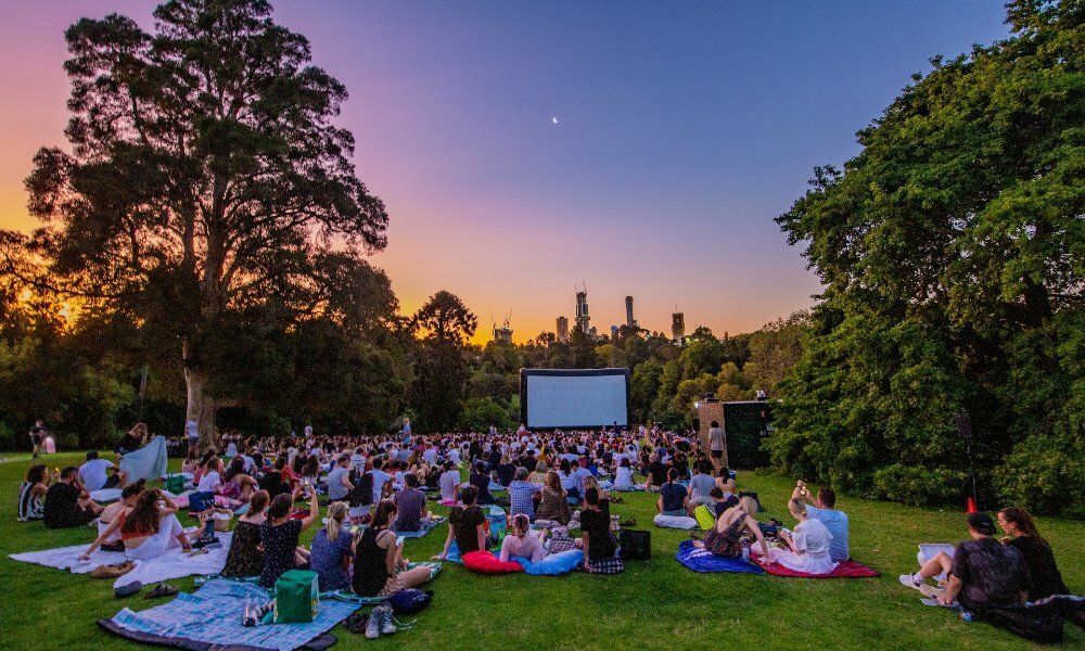 Crowds sitting on picnic blankets at an outdoor cinema at sunset.