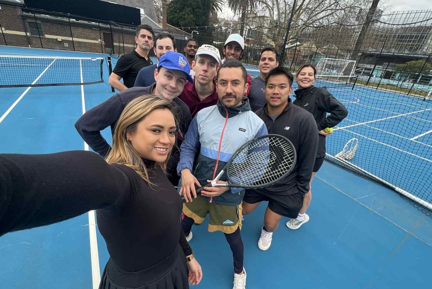 A group of smiling people standing on a tennis court, some holding racquets, taking a selfie