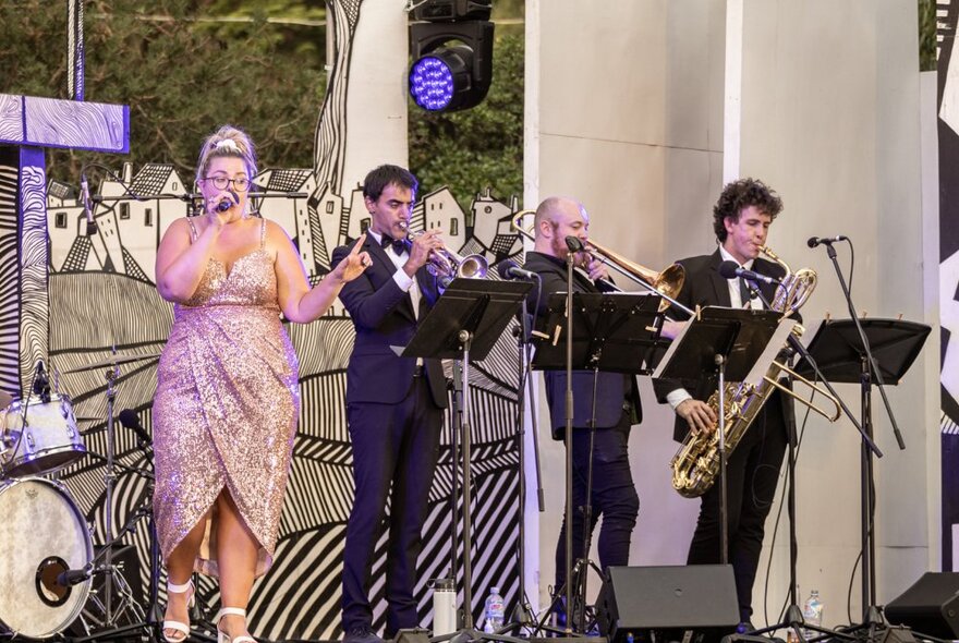 Performer Elly Poletti on stage in a shimmering sequined dress, singing into a microphone alongside three brass instrument players.