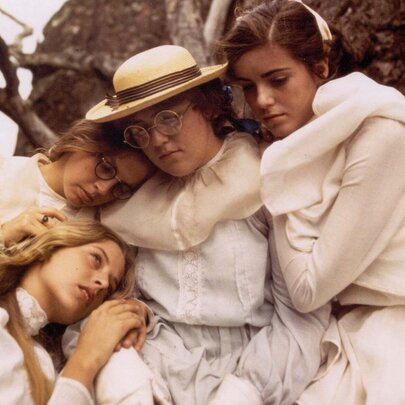 A still from the film Picnic at Hanging Rock, depicting four teenage girls wearing white Edwardian-style dresses huddling close together outdoors against a backdrop of dark, volcanic rocks.