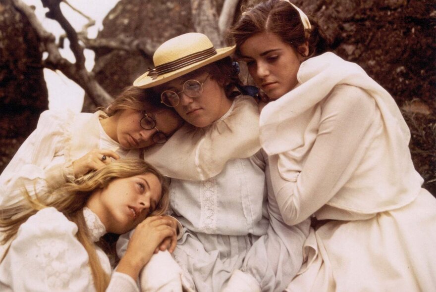 A still from the film Picnic at Hanging Rock, depicting four teenage girls wearing white Edwardian-style dresses huddling close together outdoors against a backdrop of dark, volcanic rocks.