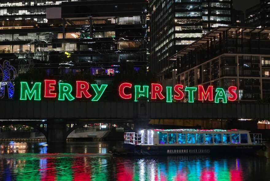 The festive 'MERRY CHRISTMAS' light display mounted on a bridge over the Yarra River at night, with water in the foreground and city buildings in the background.