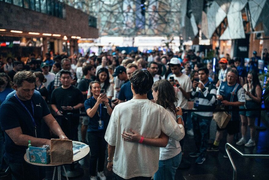 A couple with their arms around each other facing a large crowd of onlookers in the Atrium at Fed Square. 