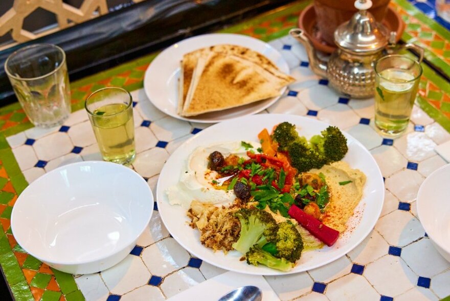 Overhead view of a vegetarian mixed plate, flat bread, and mint tea in a glass, served on a ceramic tiled mosaic table.