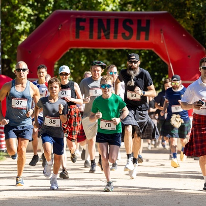 Adults and children, some wearing kilts, running through a red blow-up finish line marker during a fun run.