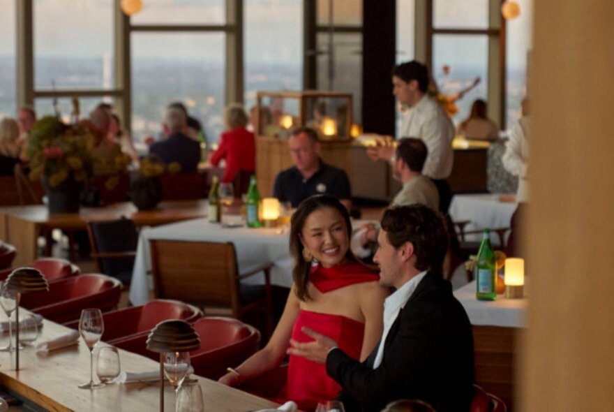 Two people dining at the bar of the restaurant Atria, in the background are white linen covered tables in a larger dining space.