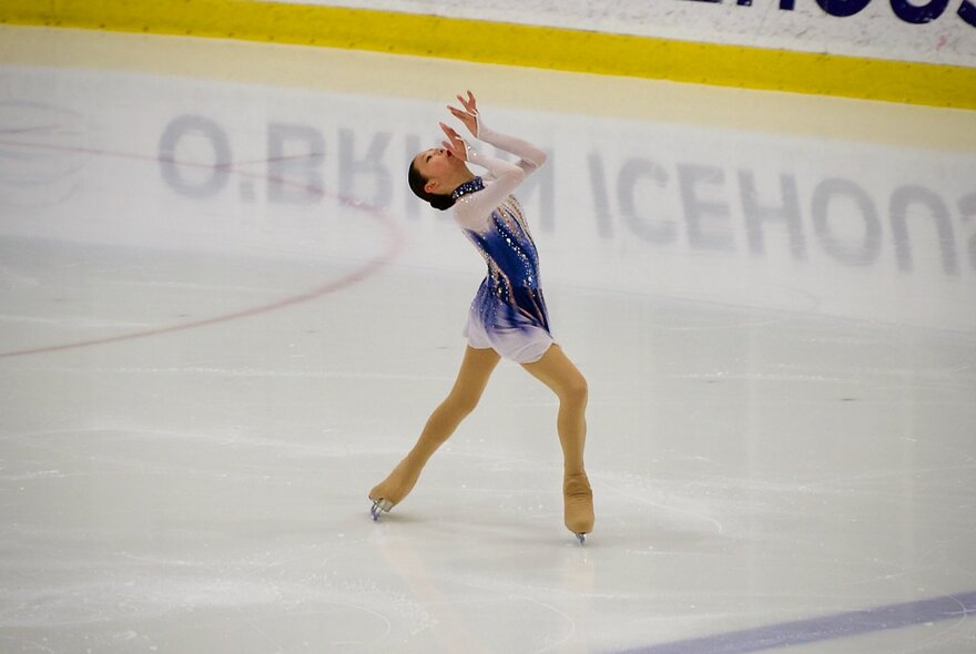 A figure skater wearing a blue and white costume skating on an ice rink, with the words O'BRIEN ICEHOUSE reflected in reverse on the ice. 