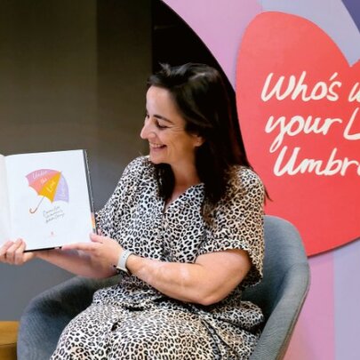 Children's author, Davina Bell, holding a book open and facing away from her as she sits in an armchair to conduct a live reading; a red heart and rainbow on the wall behind her.