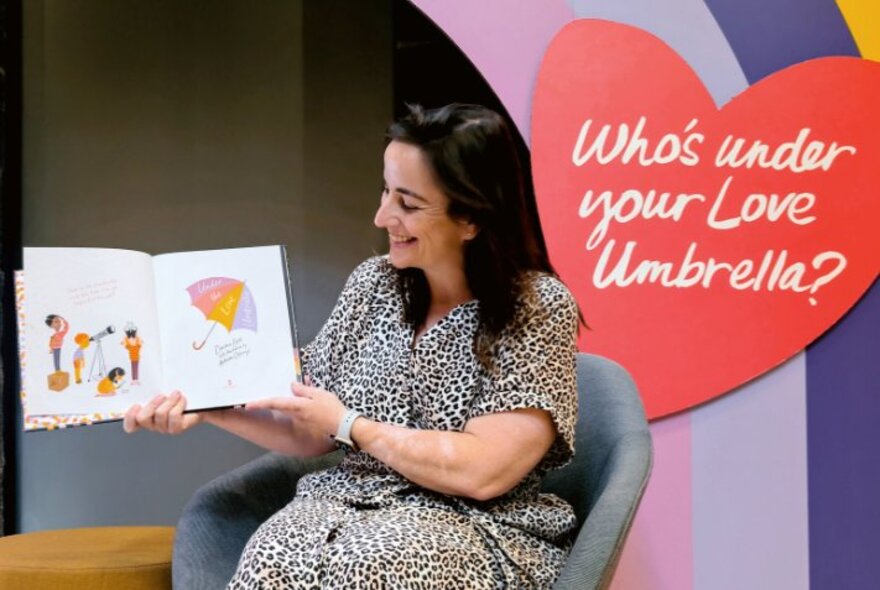 Children's author, Davina Bell, holding a book open and facing away from her as she sits in an armchair to conduct a live reading; a red heart and rainbow on the wall behind her.