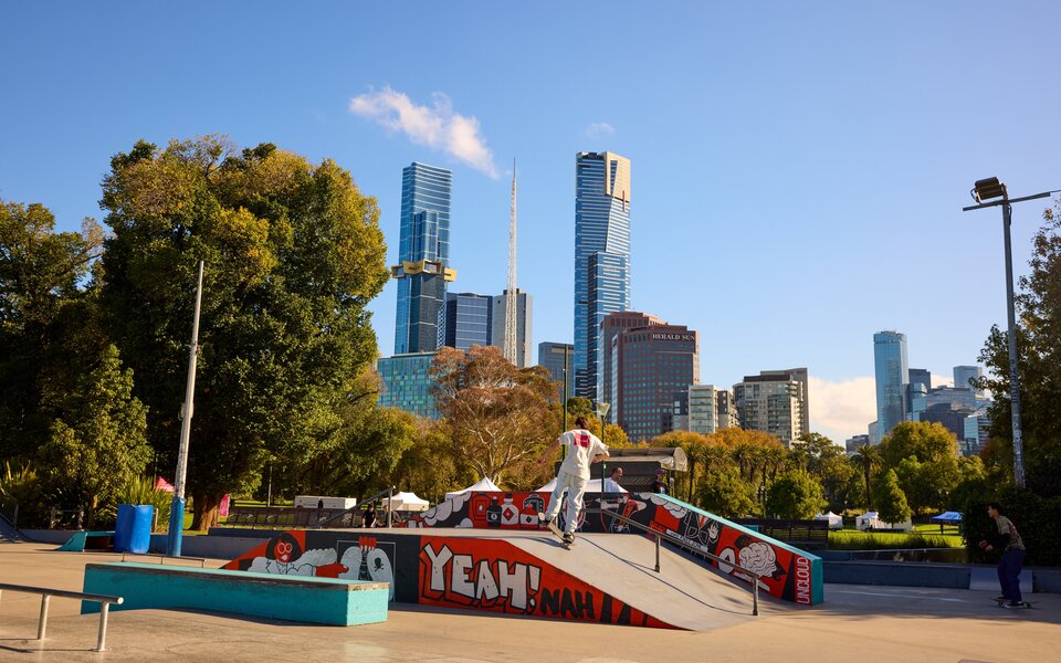 A man riding a skateboard up a ramp at a skate park with a view of the Melbourne city skyline in the background.