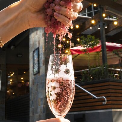A woman's hand squeezing red grapes, with the juice flowing into a champagne flute filled with sparkling wine.