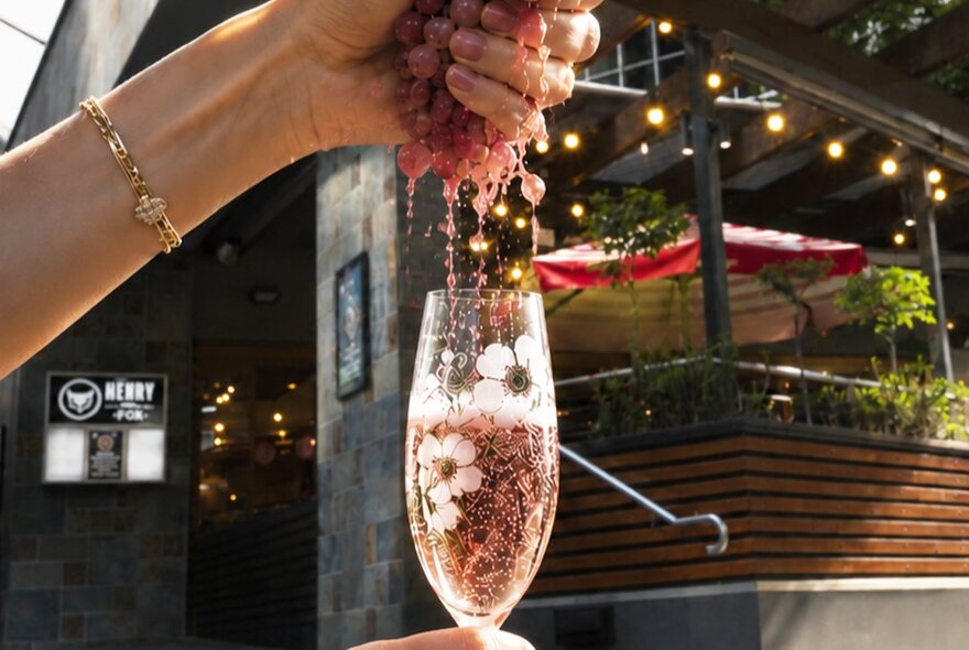 A woman's hand squeezing red grapes, with the juice flowing into a champagne flute filled with sparkling wine.