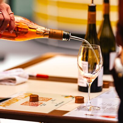 A person pouring a pale pink rosé wine from a bottle into a stemmed wine glass at a tasting event.