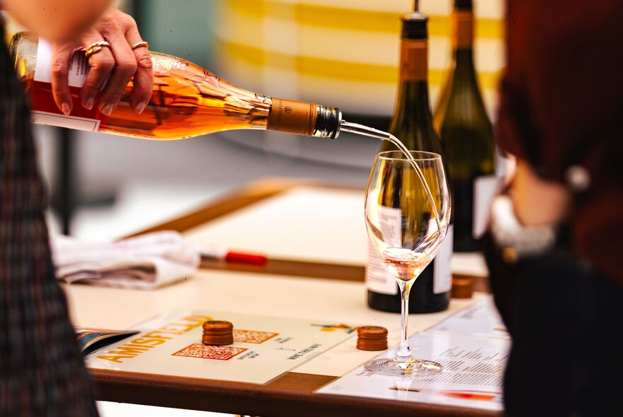 A person pouring a pale pink rosé wine from a bottle into a stemmed wine glass at a tasting event.