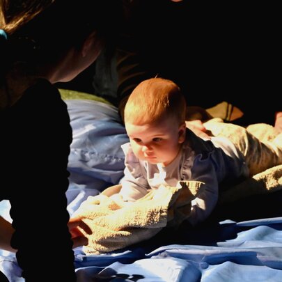 A young baby lying on its stomach on a rug, with it's carer kneeling in front.