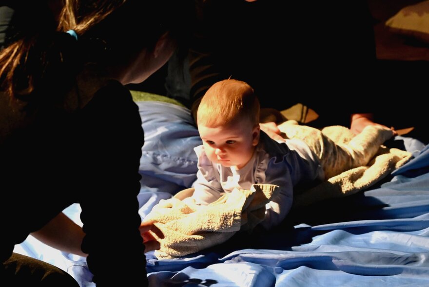 A young baby lying on its stomach on a rug, with it's carer kneeling in front.