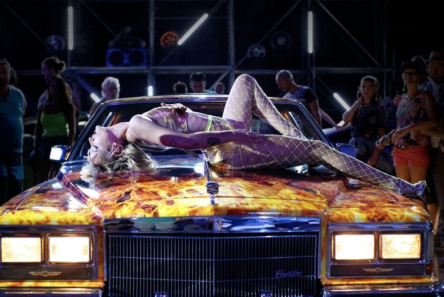 A scene from a film showing a semi-naked woman laying across a car bonnet with it's lights on. 