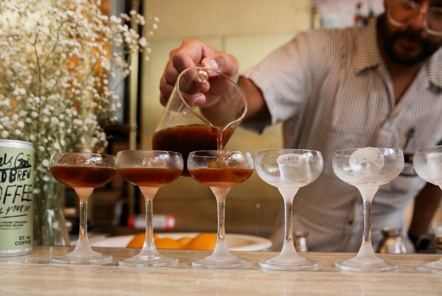 A barista pouring espresso martinis from a glass jug into six iced martini glasses.