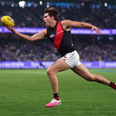 An Essendon AFL player running on the grassy field with arms outstretched reaching for the football, blurred spectators in the background.