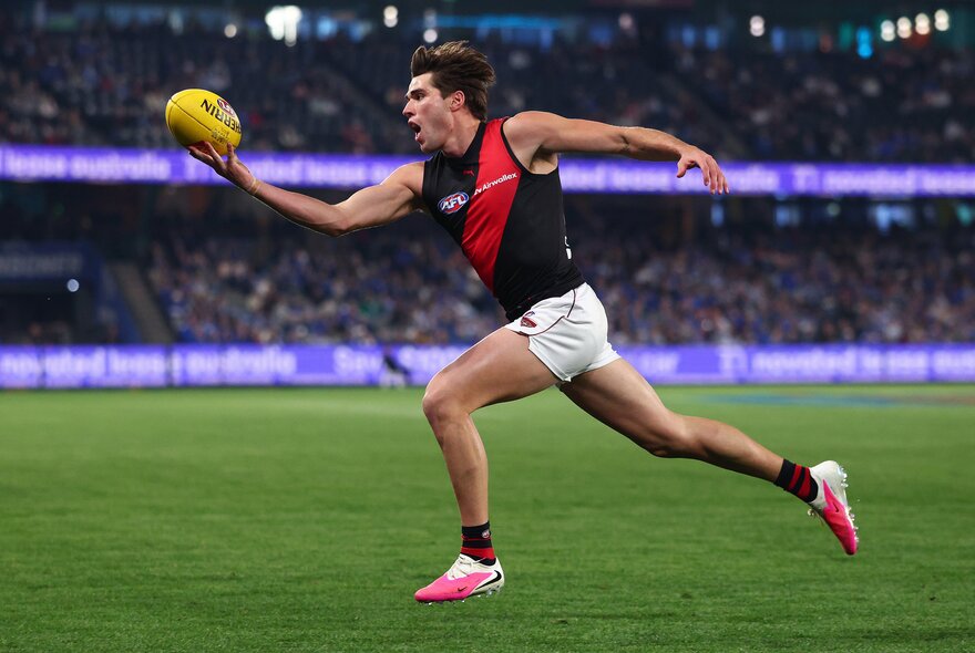 An Essendon AFL player running on the grassy field with arms outstretched reaching for the football, blurred spectators in the background.