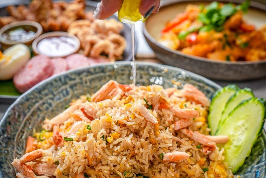 Hand squeezing lemon juice onto a Thai seafood rice dish, with other dishes in the background.