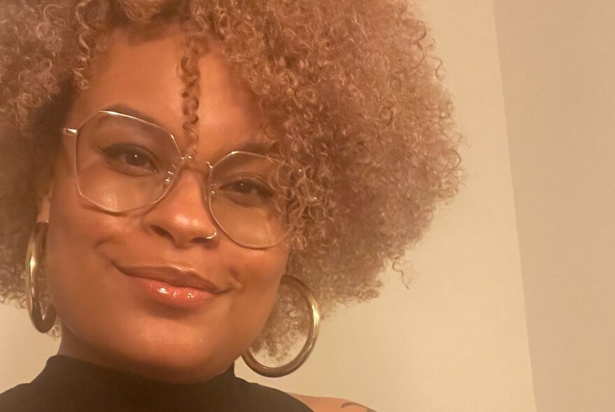 A smiling woman with tight curly hair and large glasses looking relaxed and confident.