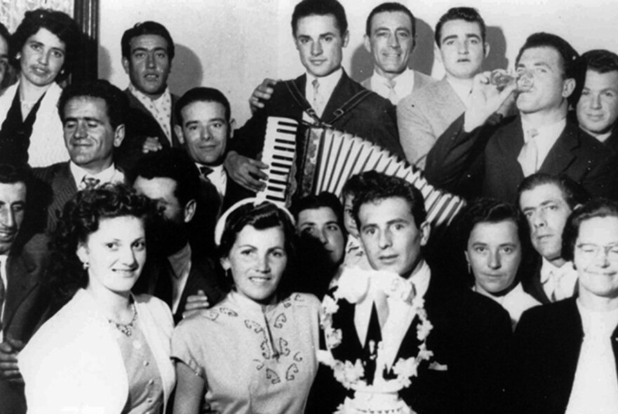 Picture from the 1950s of Italian men and women standing in a large group at a wedding, with man playing accordion.