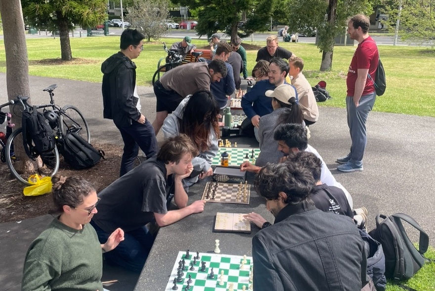 A group of people sitting in Carlton Gardens, with chess boards lined up on a concrete bench with people on either side of the bench playing games of chess.