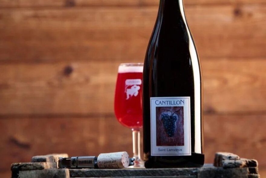 A bottle of imported beer on a table with a poured glass of red tinted beer behind it. 