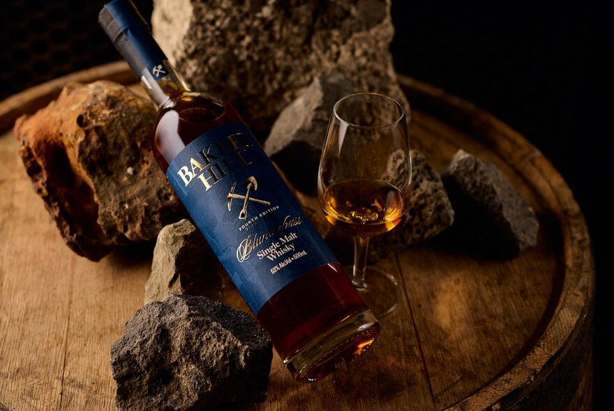 A bottle of whisky with a glass of whisky alongside, displayed against ochre coloured rocks on top of a wooden barrel.