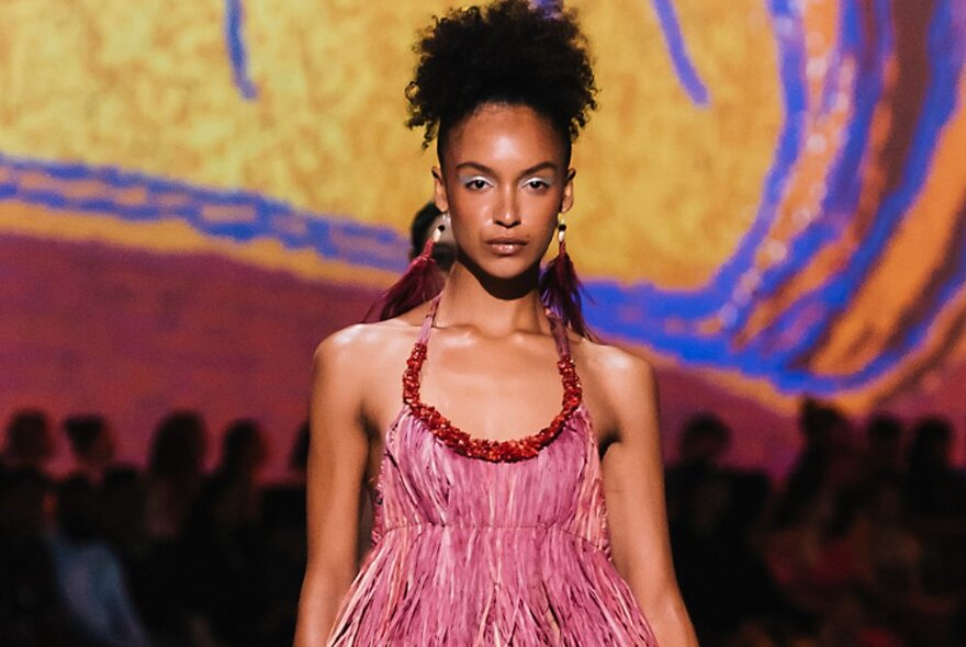 Fashion model wearing a halter neck dress and long hanging earrings walking a catwalk runway.