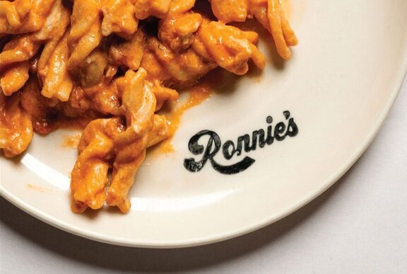 A white plate of tomato pasta, with a black logo printed on the plate.