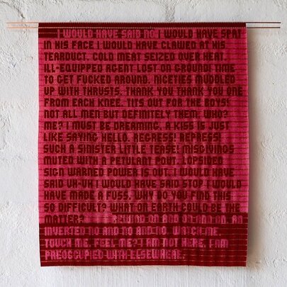 A red patterned textile artwork handing on a blank white gallery wall. 