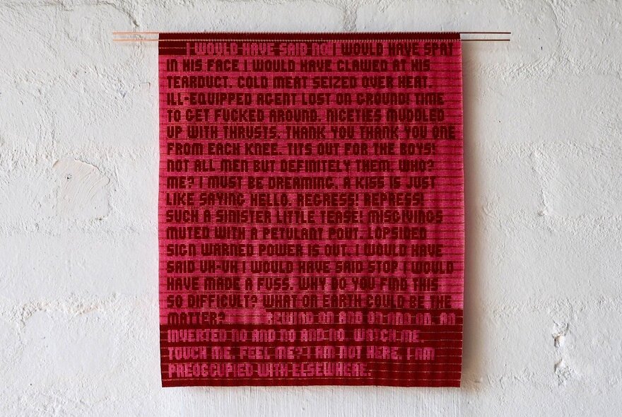A red patterned textile artwork handing on a blank white gallery wall. 
