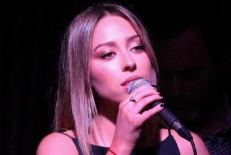 Soulful young singer Jemma Cher singing into a microphone in a pink light.