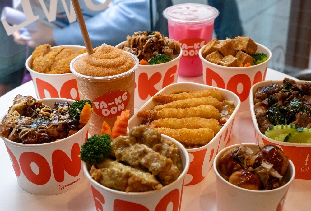 An assortment of Japanese rice bowls in takeaway containers, a side of takoyaki and two cold drinks.