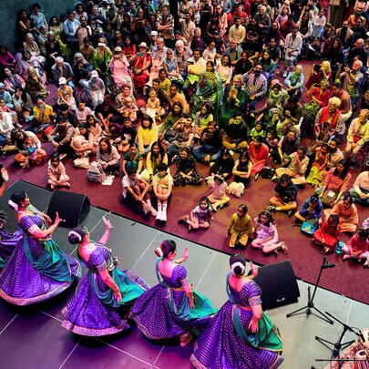 A colourfully dressed audience watching a live performance from five traditional Indan dancers, seen from above the stage.