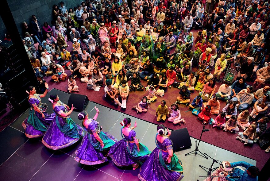 A colourfully dressed audience watching a live performance from five traditional Indan dancers, seen from above the stage.