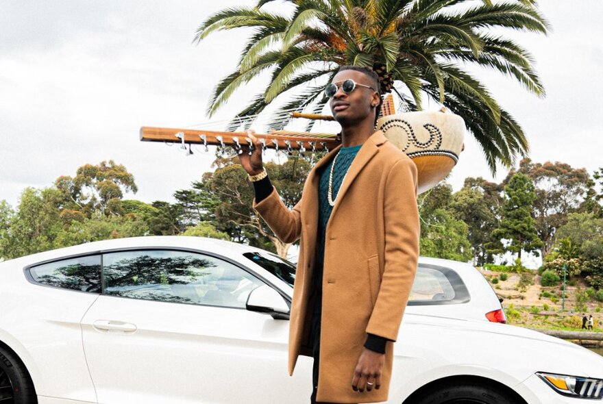 Gambian musician, Amadou Suso, holding a traditional Mandinkan instrument, the kora, over his shoulder, in front of a white sports car and palm tree.