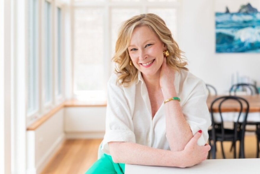 Journalist Leigh Sales sits casually leaning on a kitchen bench her one hand on her neck, smiling warmly. 