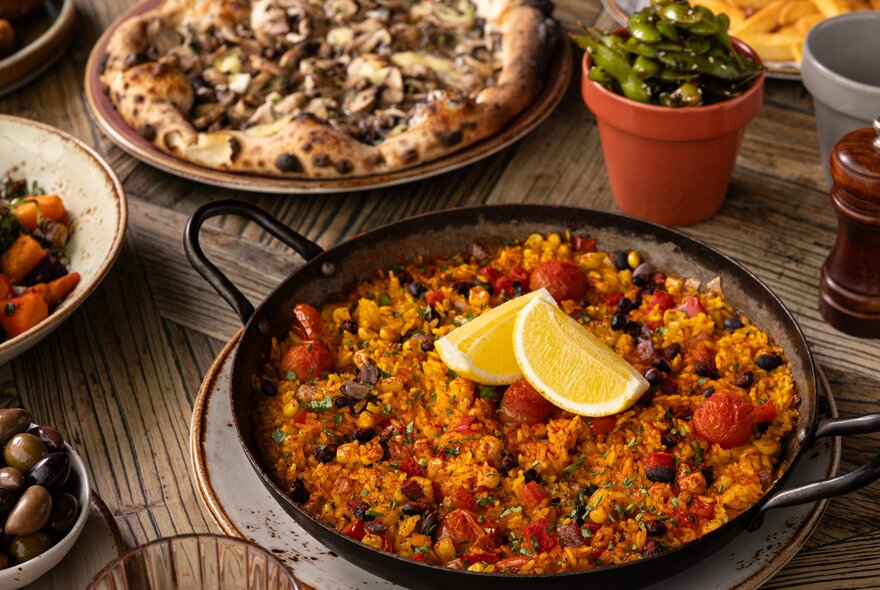 A dish of paella, and a pizza on a wooden table.