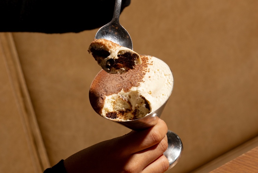 A person's hand holding a silver martini glass with tiramisu and scooping out a spoonful.