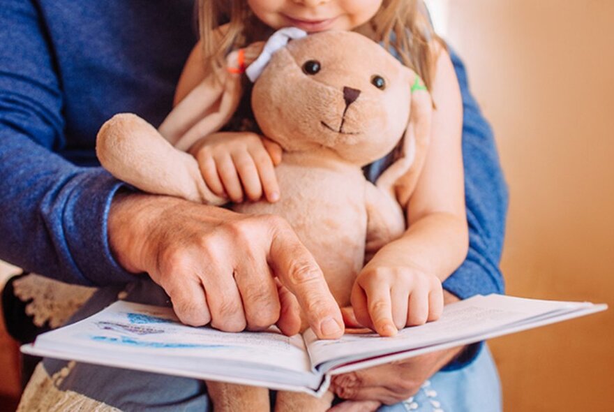 Adult with a seated child holding a teddy bear, pointing at a picture book.