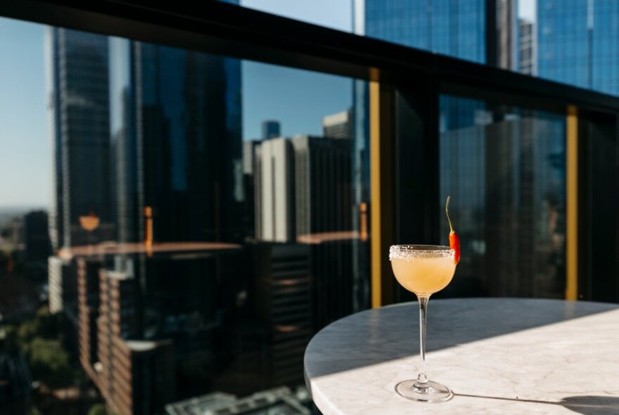 A peach-coloured cockatil resting on a round marble table on a rooftop deck, with tall city buildings behind.