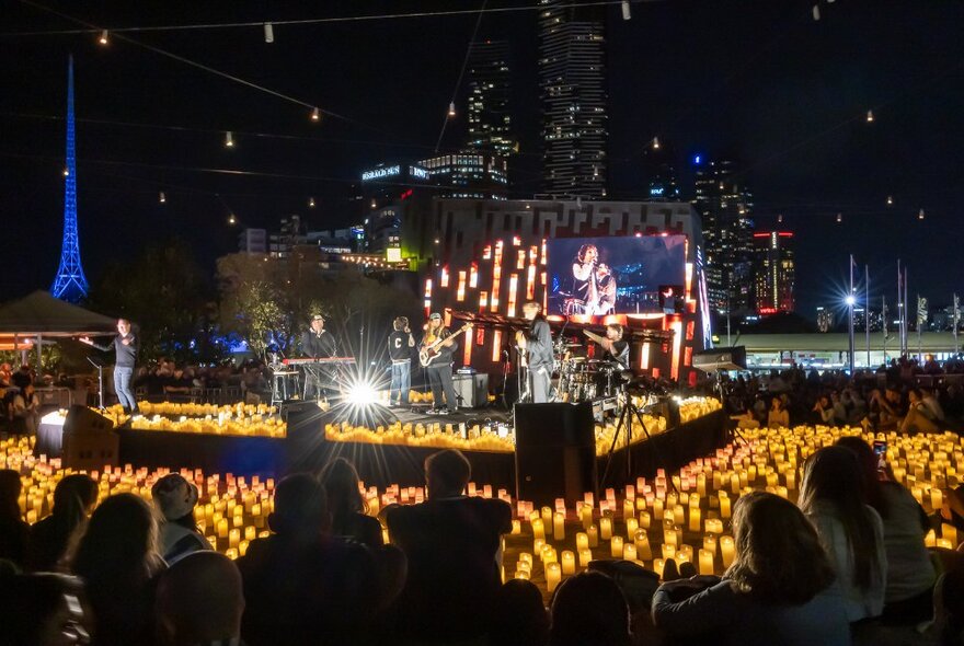 A band performing outdoors at night on a candlelit stage surrounded by a crowd.