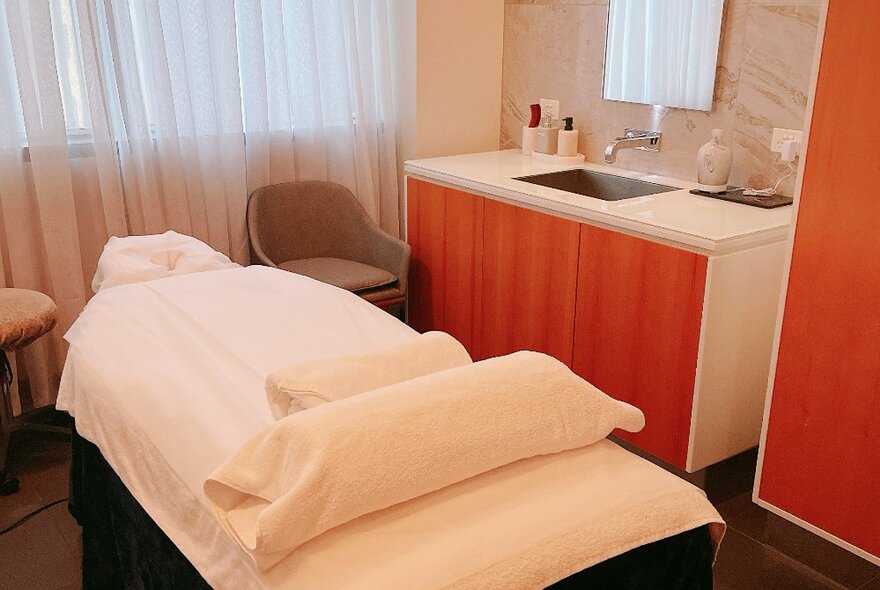 Massage table with white linen, opaque net curtains and a sink area.