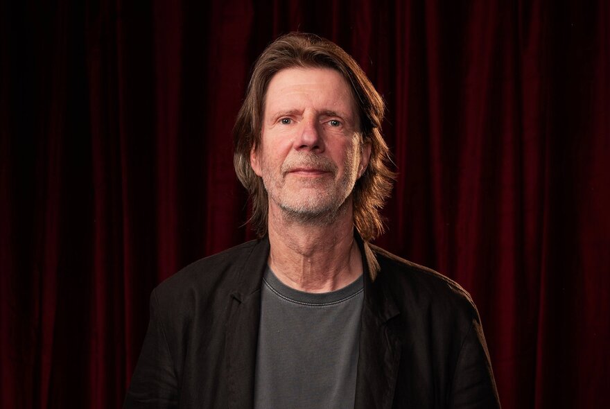 Portrait of screen composer, Burkhard Dallwitz, with wispy long hair and a composed expression on his face; a red stage curtain behind him.