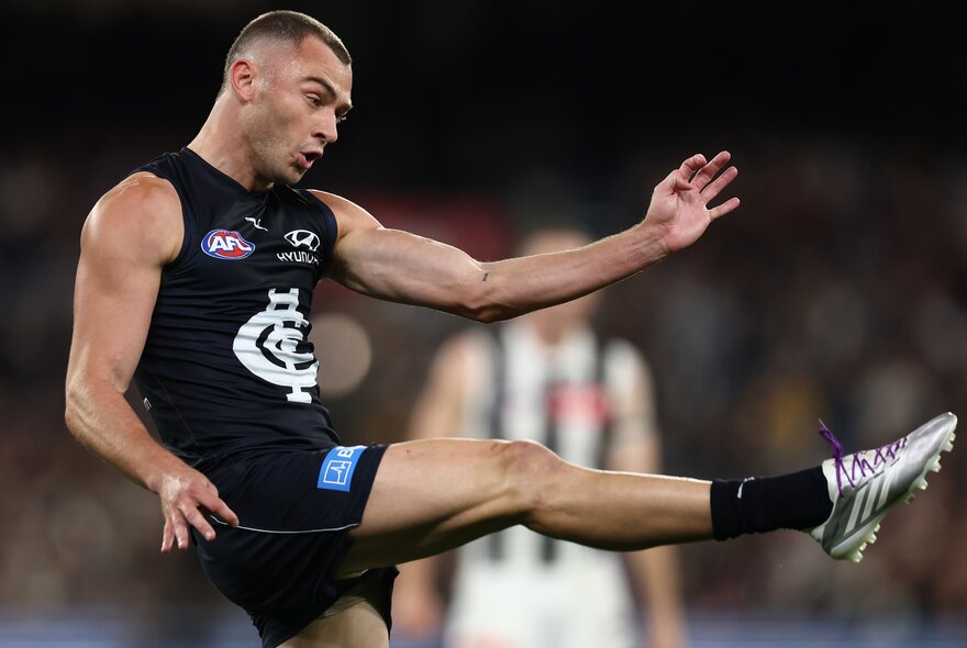 Profile of a Carlton AFL player with his leg raised high in front of him, having just kicked a football.